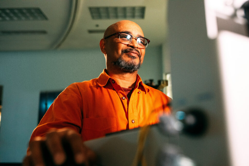 Hombre con gafas y barba, vestido con una camisa naranja, trabajando frente a una computadora
